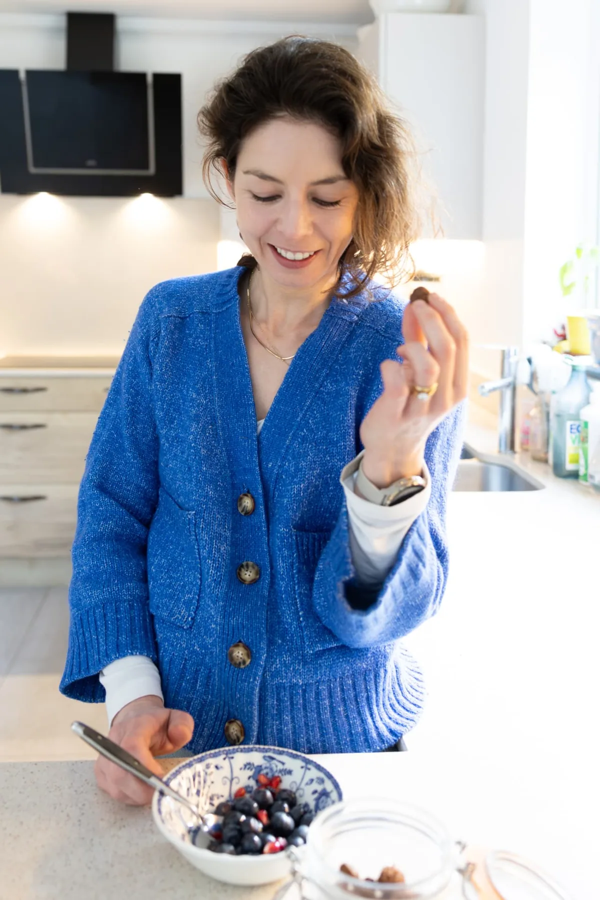 Arabella in a blue cardigan standing at her kitchen counter, holding a bowl of berries and smiling