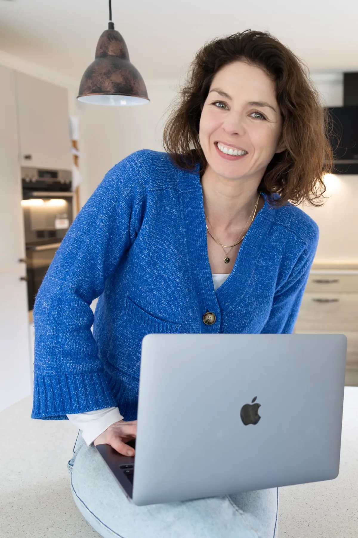 Arabella smiling in her kitchen in a blue cardigan, leaning beside an open laptop